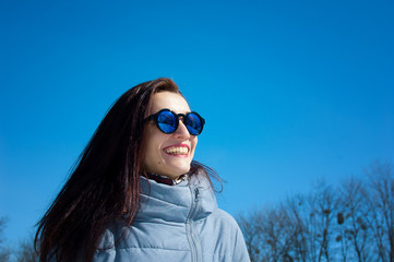 Outdoors lifestyle close up portrait of beautiful girl walking in the snowy winter park. Smiling and enjoying wintertime. Wearing stylish mirrored sunglasses, blue trench coat