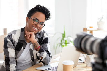 Smiling African American female blogger sit at office desk shooting video, excited black millennial...