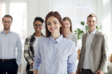 Portrait of smiling millennial female employee or worker looking at camera, happy successful woman standing foreground posing with colleagues at background, student or intern make picture in office