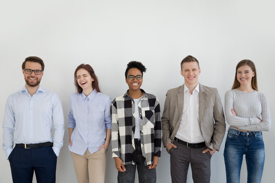 Excited multiracial millennial team standing near wall looking at camera laughing, group of young diverse employees or professionals posing, smiling business people or staff make picture in office