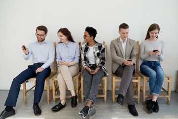 Group of multiracial young people entertain sitting in queue waiting using smartphones, diverse millennial employees communicate, chatting in line, busy with cellphones showing video or making selfie