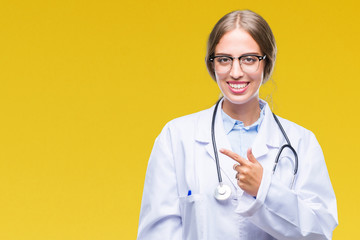 Beautiful young blonde doctor woman wearing medical uniform over isolated background cheerful with a smile of face pointing with hand and finger up to the side with happy and natural
