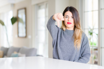 Young beautiful woman wearing winter sweater at home looking unhappy and angry showing rejection and negative with thumbs down gesture. Bad expression.