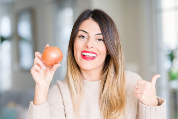 Young beautiful woman holding fresh onion at home pointing and showing with thumb up to the side with happy face smiling