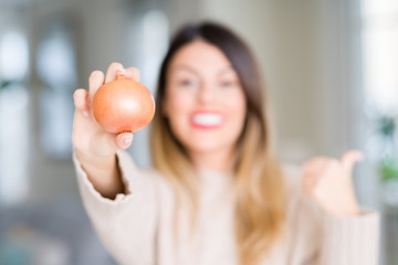 Young beautiful woman holding fresh onion at home pointing and showing with thumb up to the side with happy face smiling
