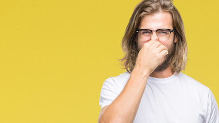 Young handsome man with long hair wearing glasses over isolated background smelling something stinky and disgusting, intolerable smell, holding breath with fingers on nose. Bad smells concept.