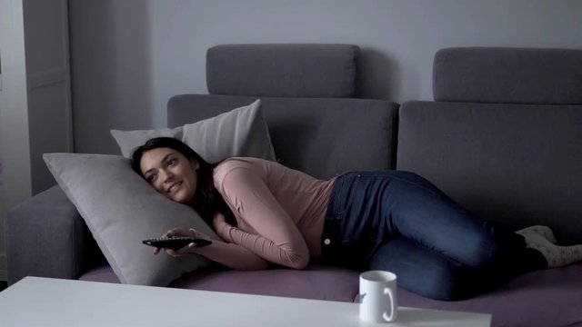 Attractive young woman lying on a sofa watching tv and laughing
