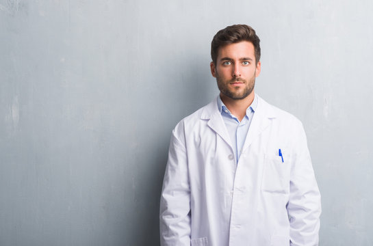 Handsome young professional man over grey grunge wall wearing white coat with serious expression on face. Simple and natural looking at the camera.