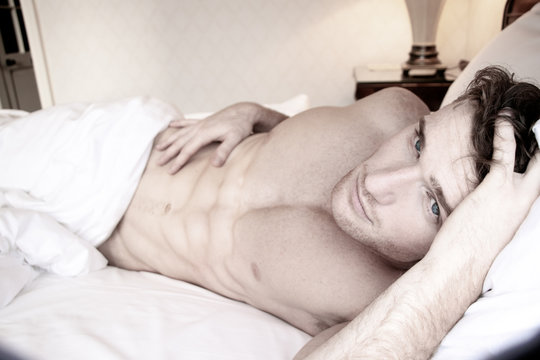 Handsome naked man with six pack abs and blue eyes lying in hotel room bed