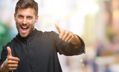 Young catholic christian priest man over isolated background approving doing positive gesture with hand, thumbs up smiling and happy for success. Looking at the camera, winner gesture.