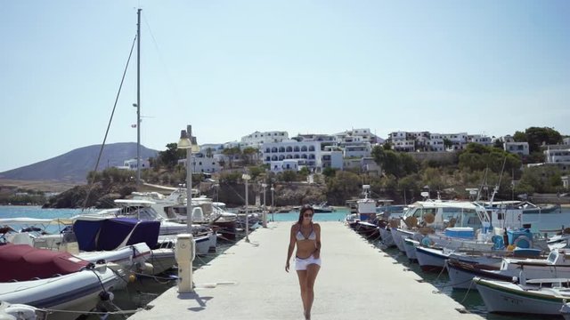 A beautiful, fit woman walks towards camera down a public pier on a warm, sunny day in Paros, Greece; white Greek homes in the background; slow motion.