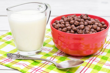 Bowl with chocolate corn balls, spoon and cup of milk on white wooden table.