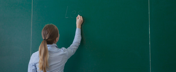 Rear view of a student or teacher with long brunette hair writing on a blank green blackboard or...