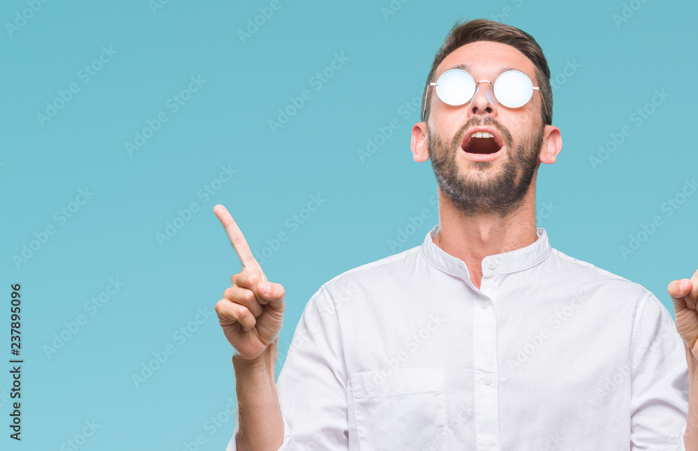 Wall mural young handsome man wearing glasses over isolated background amazed and surprised looking up and poin