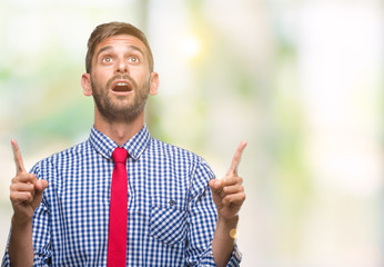 Young handsome business man over isolated background amazed and surprised looking up and pointing with fingers and raised arms.