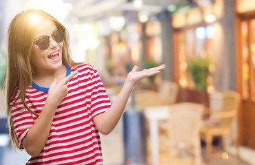 Young beautiful girl wearing sunglasses over isolated background amazed and smiling to the camera while presenting with hand and pointing with finger.