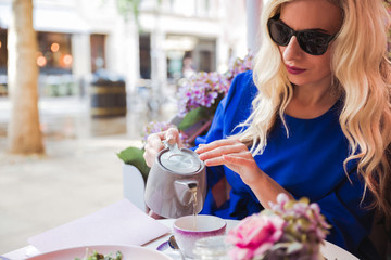 Portrait of the elegant young woman who is pouring tea from the kettle into a cup sitting at the table in the restaurant outdoor