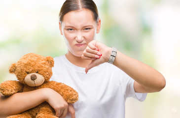 Young caucasian woman holding teddy bear over isolated background with angry face, negative sign showing dislike with thumbs down, rejection concept