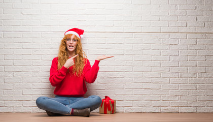 Young redhead woman sitting over brick wall wearing christmas hat amazed and smiling to the camera while presenting with hand and pointing with finger.