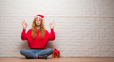 Young redhead woman sitting over brick wall wearing christmas hat relax and smiling with eyes closed doing meditation gesture with fingers. Yoga concept.