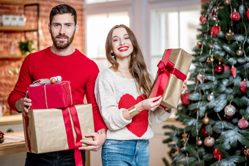 Portrait of a young lovely couple in sweaters standing together with New Year gifts near the Christmas tree at home