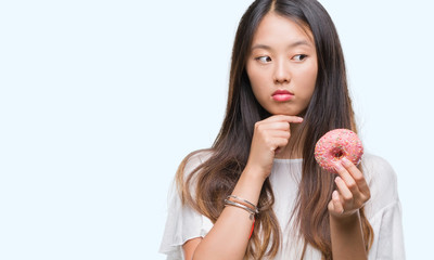 Young asian woman eating donut over isolated background serious face thinking about question, very confused idea