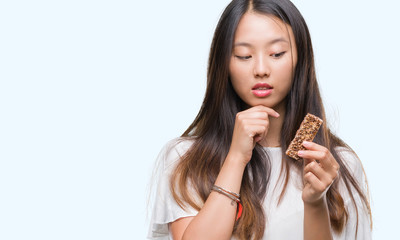 Naklejka premium Young asian woman eating chocolate energetic bar over isolated background serious face thinking about question, very confused idea