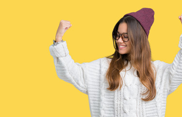 Young beautiful brunette hipster woman wearing glasses and winter hat over isolated background showing arms muscles smiling proud. Fitness concept.