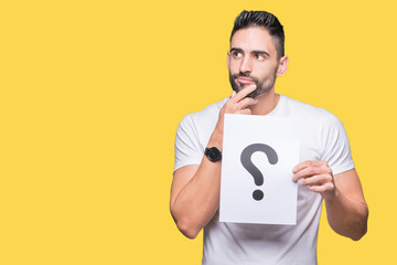 Handsome young man holding paper with question mark over isolated background serious face thinking...