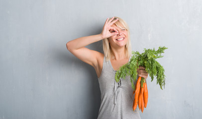 Caucasian adult woman over grey grunge wall holding fresh carrots with happy face smiling doing ok sign with hand on eye looking through fingers