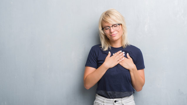 Adult caucasian woman over grunge grey wall wearing glasses smiling with hands on chest with closed eyes and grateful gesture on face. Health concept.