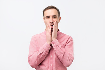 Gloomy man in pink shirt, holding hands on face, frowning and staring with helpless expression