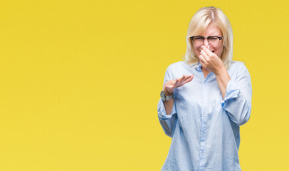 Young beautiful blonde business woman wearing glasses over isolated background smelling something stinky and disgusting, intolerable smell, holding breath with fingers on nose. Bad smells concept.