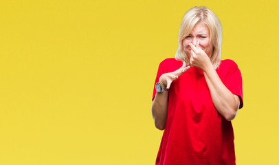 Young beautiful blonde woman wearing red t-shirt over isolated background smelling something stinky and disgusting, intolerable smell, holding breath with fingers on nose. Bad smells concept. - obrazy, fototapety, plakaty