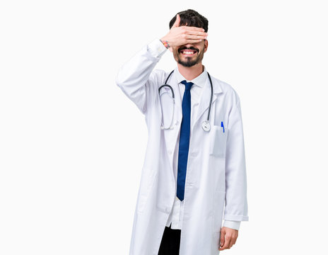 Young doctor man wearing hospital coat over isolated background smiling and laughing with hand on face covering eyes for surprise. Blind concept.