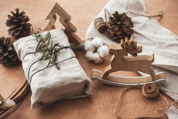Stylish christmas rustic gift wrapped in linen fabric with green branch on wooden table with pine cones, tree,reindeer, twine, cotton. Simple eco presents plastic free. Zero waste  holidays.