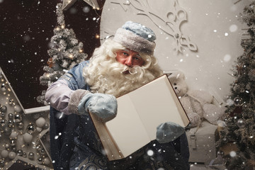 Russian Santa Claus (Grandfather Frost) in a blue caftan sits and holds in his hands a large open book with empty pages for wishes. - obrazy, fototapety, plakaty