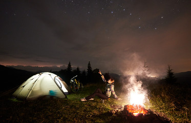 Young woman cyclist having a rest at night camping, pointing at evening sky full of stars, sitting near burning campfire, illuminated tourist tent, mountain bike. Outdoor activity and tourism concept