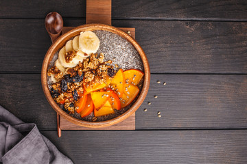 Chia pudding with banana, persimmon and granola, dark wooden background, top view, copy space.
