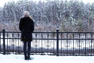 a girl in a black coat stands with her back alone on the waterfront near the forest in winter