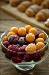 Frozen cherries and cherry plums in a glass dish against the background of fresh baked croissants on a wooden stand.