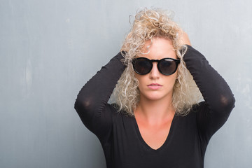 Young blonde woman with curly hair over grunge grey background suffering from headache desperate and stressed because pain and migraine. Hands on head.