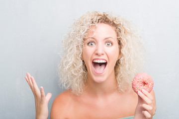Young blonde woman over grunge grey background eating pink donut very happy and excited, winner expression celebrating victory screaming with big smile and raised hands