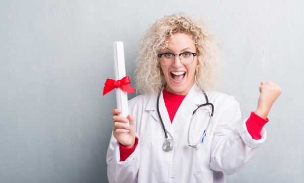 Young blonde doctor woman over grunge grey wall holding degree screaming proud and celebrating victory and success very excited, cheering emotion