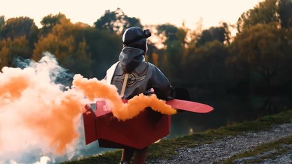 Camera follows happy little aviator boy running along sunset lake in fun cardboard plane with color smoke slow motion.