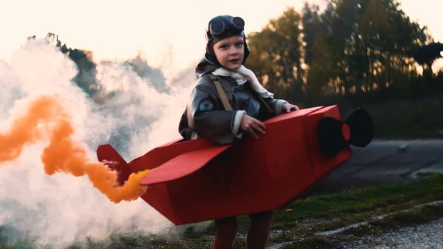 Happy little aviator boy running in circles wearing fun cardboard plane pilot costume with red color smoke slow motion.