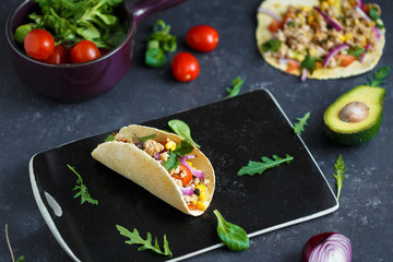 Mexican tacos with pork, vegetables, tomatoes, avocado and spices on a black stone plate on a dark background with ingredients for tacos