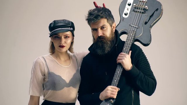 Bearded man with a guitar and a woman in a leather hat are looking at each other. Hard rock couple with guitar. Woman wearing a leather hat makes horns by bearded man with a guitar.