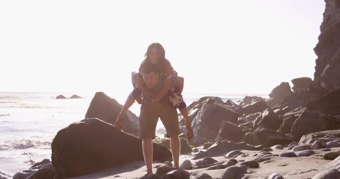 Young couple being silly and having fun at beach during sunset - woman jumps down from ontop of mans back
