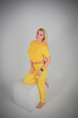 A girl in full growth in a yellow shirt and yellow pants looking into the camera with her knee on a white cube.